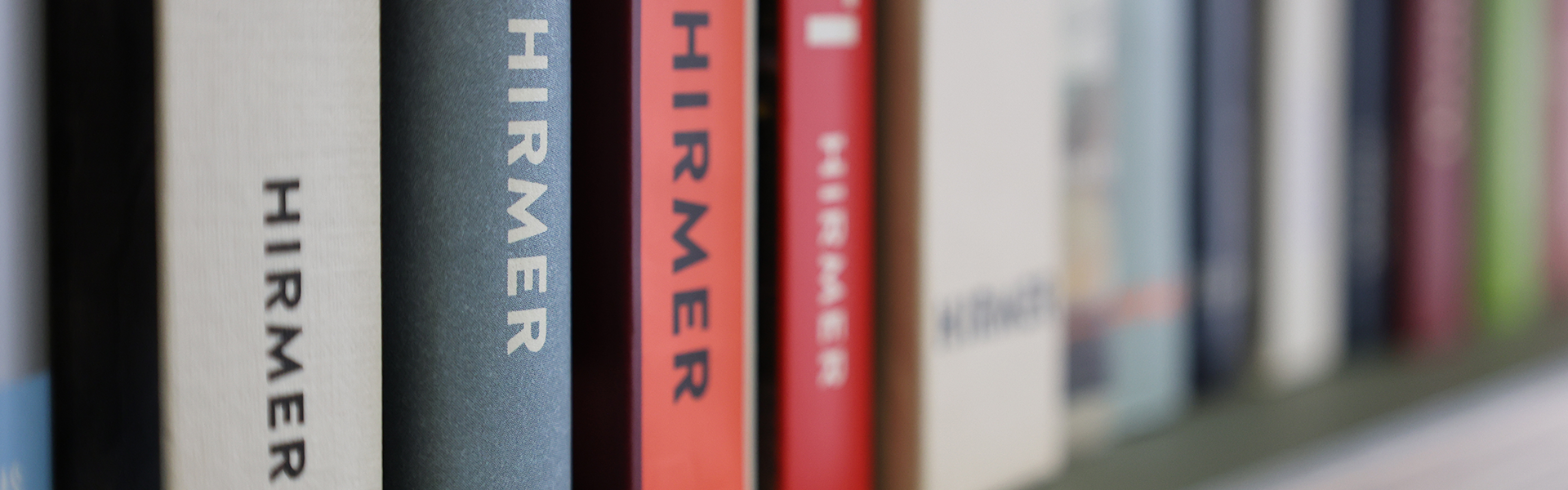 Nahaufnahme von Büchern, die aufrecht in einem Regal stehen und auf deren Rücken in verschiedenen Farben das Wort HIRMER senkrecht geschrieben ist. Der Fokus liegt auf den Buchrücken, wobei einige scharf und andere unscharf abgebildet sind.